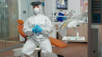 Dentistry doctor wearing protection suit speaking on camera with patients during pandemic coronavirus. Orthodontic on call with coverall, face shield, mask, gloves with assistant in background video