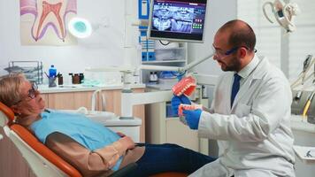 Young stomatologist explaining to senior patient procedure using model of dental teeth. Doctor holding sample of human jaw telling informations for keeping healthy teeth, digital x-ray in background video