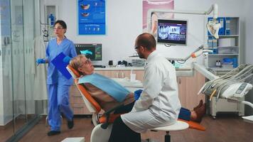 ortodontista discutindo mri Varredura com paciente sentado dentro estomatológico cadeira dentro dental clínica. idosos mulher explicando dental problema para médico indicando boca enquanto enfermeira trabalhando dentro fundo video