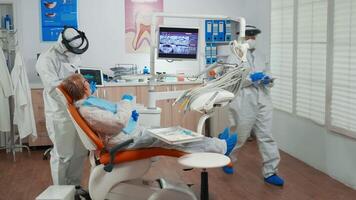 Dentist in protective equipment showing on tablet dental x-ray reviewing it with senior patient. Medical team wearing face shield coverall, mask, gloves, explaining radiography using notebook display video