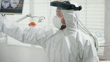 Portrait of dentist with face shield explaining holding dental x-ray to patient pointing on monitor during covid-19 pandemic. Orthodontic doctor wearing protection suit, coverall, mask and gloves video