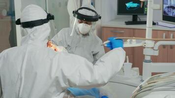 Dentists with face shield cleaning and examining patient teeth in stomatological clinic during global pandemic. Assistant and orthodontic doctor wearing protection suit, coverall, mask and gloves. video