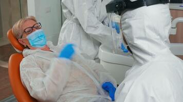 Patient explaining dental problem to stomatologist wearing protection suit during global pandemic. Assistant and doctor with coverall face shield mask gloves examining woman from stomatological chair video