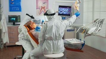 Nurse and doctor in protection suit working in dental unit during coronavirus pandemic treating senior patient. Assistant and orthodontic doctor wearing coverall, face shield, mask and gloves. video