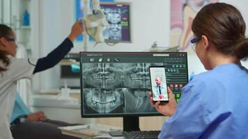 Nurse having call with expert stomatologic medic while doctor is working with patient in background. Stomatologist assistant listening dentist using mobile webcam sitting on stomatological chair video