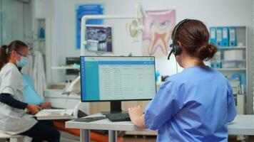 dentista assistente fabbricazione appuntamenti utilizzando cuffia seduta nel davanti su computer mentre medico è Lavorando con paziente nel sfondo l'esame denti problema. infermiera assunzione Appunti nel stomatologiche ufficio video