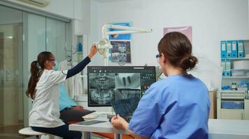 Stomatologist nurse comparing radiographics looking at computer, while specialist doctor with face mask speaks to man with toothache sitting on stomatological chair preparing tools for surgery video