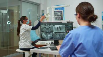 Nurse analysing digital x-ray sitting in front of pc in stomatologic clinic, while doctor with face mask is working with patient in background examining teeth problems, lighting the lamp video