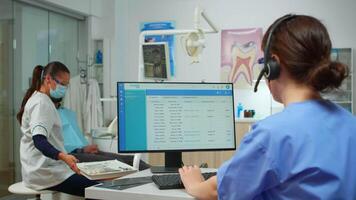 stomatologo infermiera parlando con pazienti utilizzando cuffia fabbricazione dentale appuntamenti seduta nel davanti su computer mentre medico è Lavorando con paziente sfondo l'esame denti problema video