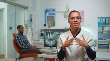 Stomatologist doctor talking on web cam with patients about oral hygiene looking at camera while patient waiting for nurse in background. Dental assistant preparing man for stomatological surgery. video