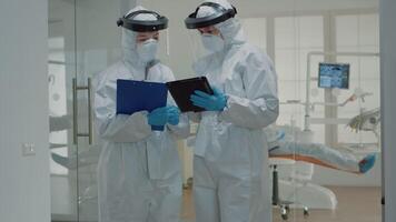 Dentist giving tablet to dental nurse before consultation appointment with patient. Stomatology doctors wearing ppe suits during pandemic while treating man for oral healthcare video