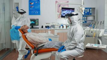 Dentist nurse in coverall putting dental bib to old woman before stomatological examination during covid-19 pandemic. Assistant and orthodontic doctor wearing protection suit, face shield mask gloves video