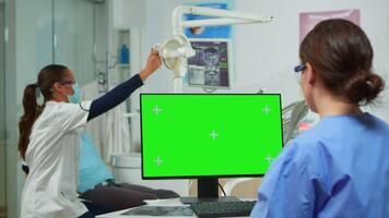 dentiste infirmière à la recherche à vert écran afficher tandis que médecin examens patient mensonge sur dentaire chaise. stomatologue assistant en utilisant moniteur avec vert chrominance clé isolé afficher maquette écran tactile video