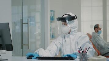 Nurse wearing ppe suit sitting at oral care clinic desk and using professional computer. Dentistry assistant answering telephone for teethcare appointments while typing on keyboard video