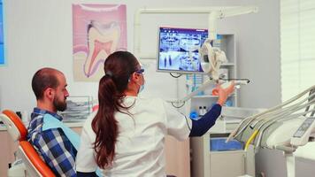 Stomatologist doctor lighting the lamp preparing patient for dental surgery. Dentist with face mask examining patient with dental bib before dental intervention sitting on stomatological chair video