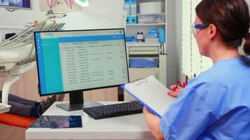 Orthodontist nurse looking at list with appointments in computer and taking notes on clipboard while dentistry doctor is working with patient background examining teeth problems.. video