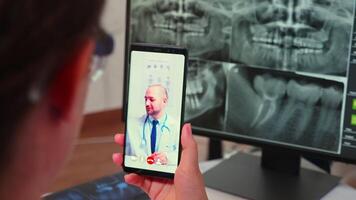 Close up of assistant having videocall with specialist stomatologist using smartphone sitting in modern dental clinic in front of pc with digital x-ray. Dentistry doctor explaining patient symptoms video