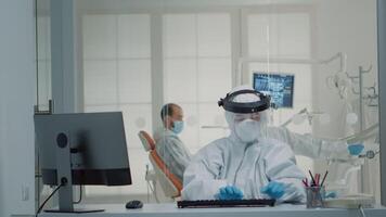 Stomatology assistant with ppe suit using computer while sitting at oral clinic desk. Nurse talking on office telephone while professional dentist consulting patient in background video