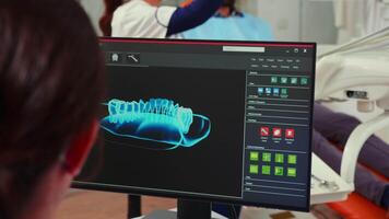 Stomatologist nurse looking at digital dental fingerprint on computer sitting in stomatological clinic while dentistry doctor is working with patient in background examining teeth problems. video