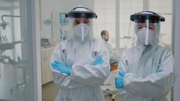 Professional team of dentists in ppe suits standing in dental cabinet looking at camera. Stomatologists wearing virus protection uniform preparing for oral patient examination video