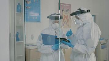 Stomatologists wearing hazmat suits using tablet for dental hygiene and consultation. Dentist and assistant doing patient examination with professional equipment during pandemic video