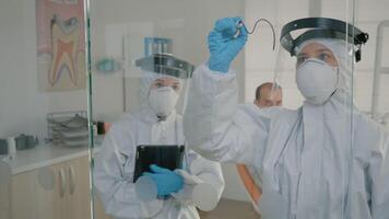 equipo de estomatólogos vistiendo ppe trajes utilizando tableta y negro marcador. dental especialista dibujo diente modelo para referencia en estomatología gabinete vaso. ortodoncistas operando durante pandemia video