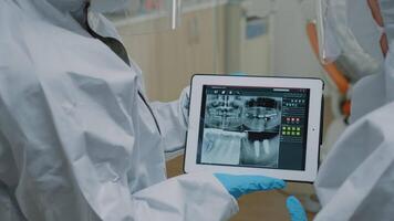 Close up of teeth radiography scan on modern tablet while dental specialist holding device and assistant looking at screen. Stomatologists wearing ppe suits examining oral x ray video