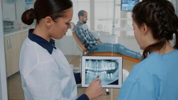 Stomatologist holding modern tablet with x ray on screen examining oral care and hygiene. Nurse and dentist looking at radiography discussing teeth diagnosis for patient sitting in cabinet video