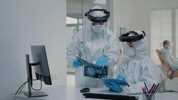Orthodontist and assistant using x ray of teeth for oral care, wearing virus protection suits at clinic. Stomatologists looking at computer on desk and dental radiography in hand video