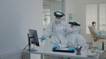 Stomatology team of dentists at desk using computer for teethcare in dental cabinet, wearing virus protection suits. Nurse with modern device sitting while orthodontist preparing for patient video