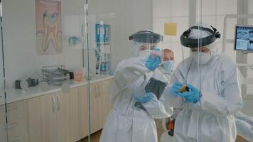 Dentistry team of specialists using sticky notes on glass in stomatology cabinet to explain teethcare diagnosis to patient. Dentist and nurse wearing ppe suits for examination during pandemic video