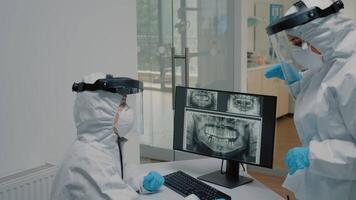 Dentistry staff looking at dental x ray scan wearing ppe suits against virus pandemic at stomatology cabinet. Orthodontist and assistant using computer screen and radiography technology video