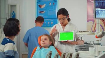 dentiste montrant vert écran afficher à mère de peu patient tandis que fille mensonge sur dentaire chaise. femme expliquant en utilisant moniteur avec vert chrominance clé isolé chrominance PC clé maquette écran vert video