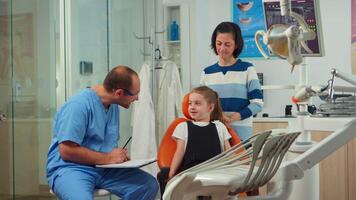 amigáveis homem dentista assistente interrogando criança paciente e levando notas em prancheta esperando para doutor. mãe do pequeno menina explicando dental problema para enfermeira indicando às dor de dente problemas. video