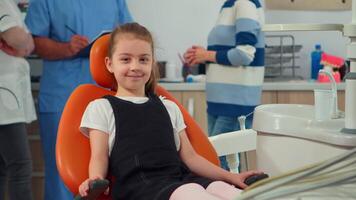 retrato do pequeno menina paciente olhando às Câmera esperando para pediatra estomatologista dentro dental clínica. criança deitado em estomatológico cadeira sorridente às Webcam enquanto enfermeira e médico falando com mãe. video