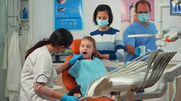 stomatologue médecin vérification dent santé de Jeune patient en utilisant dentaire outils travail dans moderne stomatologique Bureau avec homme infirmière. mère et fille visite médecin pour périodique consultant video