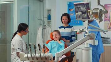 Pediatric woman dentist talking with mother and girl about eating habit and oral hygiene of the child before dental examination. Sad kid patient is showing sick tooth while nurse preparing dental tools video