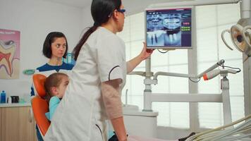 pediatra dentista mostrando em monitor dentes raio X revendo isto com mãe e filha. médico e enfermeira trabalhando juntos dentro moderno estomatológico clínica, explicando para velho mulher radiografia do dente video