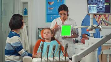 Pediatric dentist standing near girl patient pointing at green screen display while sitting on dental chair. Stomatologist explaining to mother using monitor with green chroma key isolated chroma pc video