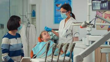 Stomatologist talking with mother of kids, lighting the lamp and examining little girl standing near stomatological chair. Pediatric dentist speaking to woman while nurse preparing sterilized tools. video