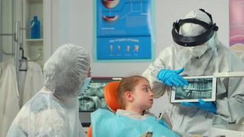 Dentist in protective equipment showing on tablet dental x-ray reviewing it with mother of kid patient. Medical team wearing face shield coverall, mask, gloves, explaining radiography using notebook video