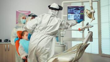 Dentist in ppe suit pointing on digital dental monitor explaining x-ray to mother and kid wearing coverall during global pandemic. New normal dentist visit in coronavirus outbreak with face shield video