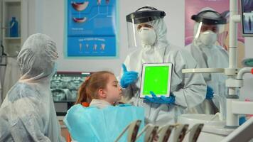 Dentist with face shield pointing at green screen display talking to child with toothache during coronavirus epidemic. Explaining using monitor with chroma key izolated chroma pc key mockup display video