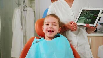 fechar acima do feliz menina paciente olhando às Câmera rindo, esperando para pediatra estomatologista dentro dental unidade. criança deitado em estomatológico cadeira sorridente às Webcam enquanto médico falando com mãe video