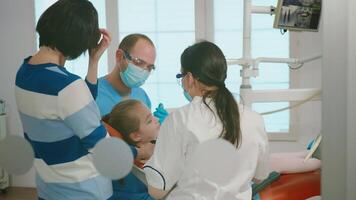 odontología doctores tratando dental problemas de niño, madre en pie cerca estomatológico silla hablando con muchacha. dentista con guantes y máscara elimina diente decaer perforación dientes durante cirugía video