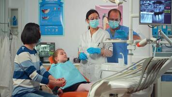 pädiatrisch Zahnarzt mit Maske Überprüfung Dental Gesundheit von ein wenig Mädchen Sitzung im stomatologisch Stuhl, Arzt mit sterilisiert Dental Instrumente, Arbeiten mit Mann Krankenschwester im modern stomatologisch Einheit. video