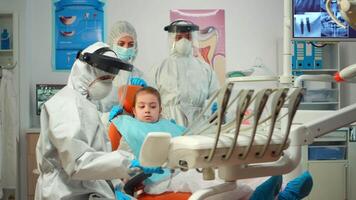 Pediatric dentist in protective suit lighting the lamp until examination while little patient opening mouth. Medical team talking wearing face shield, coverall suit, mask and gloves during coronavirus video