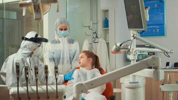 odontología enfermero en bata poniendo dental babero a niño antes de estomatológico examen durante covid-19 pandemia. concepto de nuevo normal dentista visitar en coronavirus brote vistiendo protector traje video