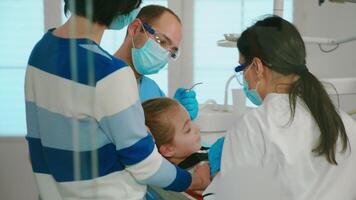 Dentists with gloves and mask removes tooth decay drilling teeth during surgery. Dentistry doctors treating dental problems of child, mother standing near stomatological chair talking with girl. video