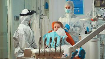 Child in protection suit showing dental problem to orthodontist wearing coverall during global pandemic. Concept of new normal dentist visit in coronavirus outbreak wearing face shield video